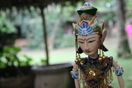 A traditional puppet featuring intricate designs and bright colors is positioned with a blurred natural background. The puppet has detailed facial features and is adorned with beaded decorations and an ornate headdress. The setting appears outdoors with soft lighting suggesting a serene environment.