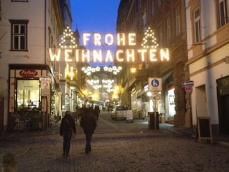 A cobblestone street in a European city is illuminated with festive holiday lights displaying the words 'Frohe Weihnachten'. The street is lined with shops and buildings, and a few people are walking, adding a bustling yet cozy atmosphere.
