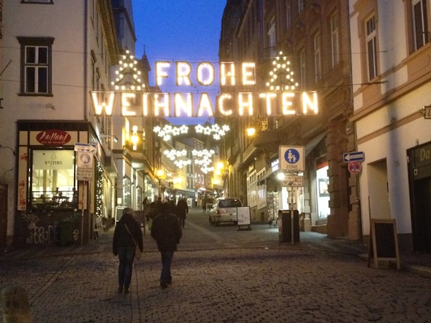 A cobblestone street in a European city is illuminated with festive holiday lights displaying the words 'Frohe Weihnachten'. The street is lined with shops and buildings, and a few people are walking, adding a bustling yet cozy atmosphere.