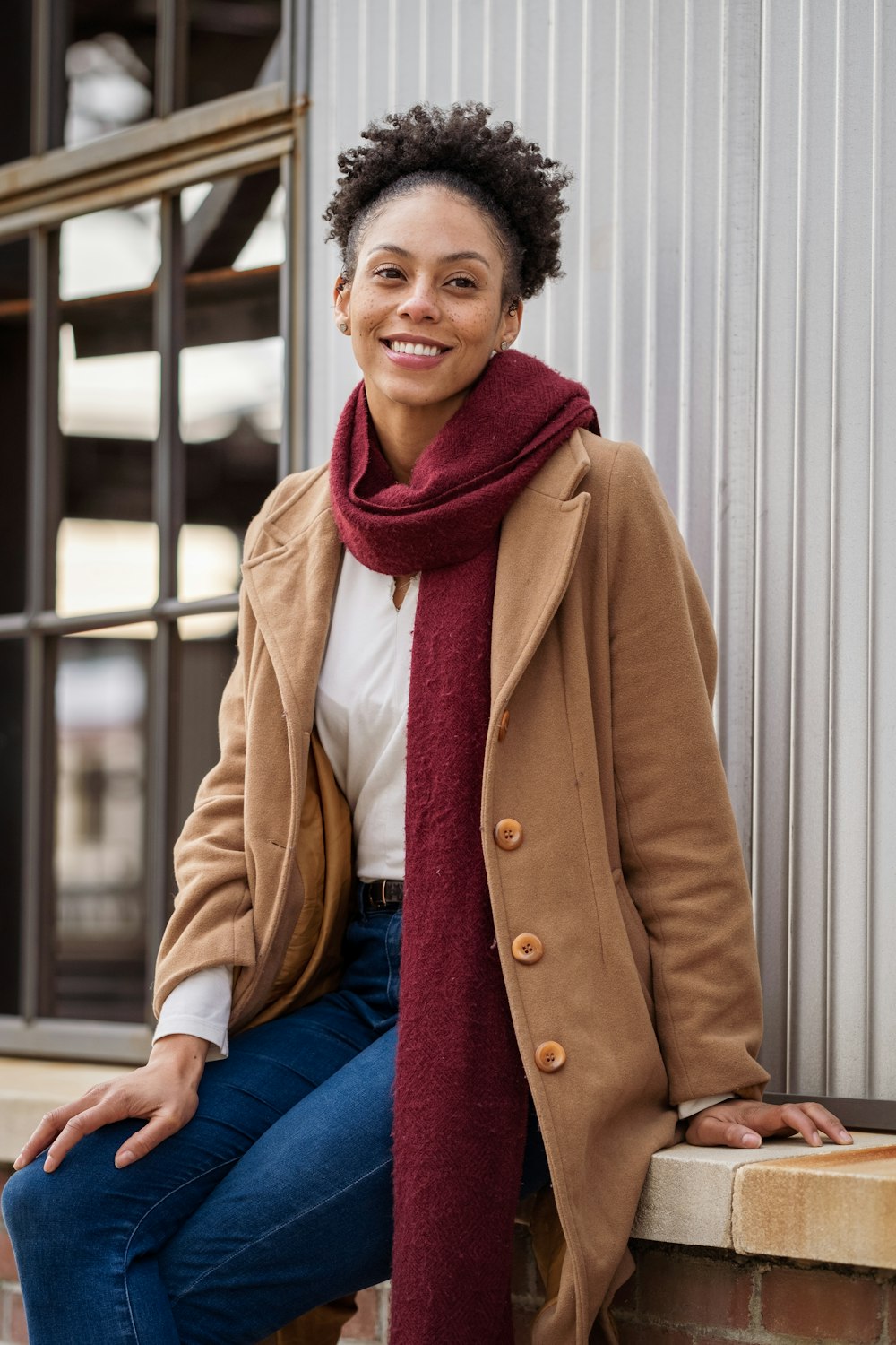smiling woman in brown coat and blue denim jeans sitting on white wooden bench