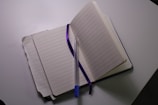 An open notebook with lined pages is placed on a flat surface. A blue pen rests diagonally across the notebook, and a purple ribbon bookmark is laid between the pages. The notebook is opened wide, revealing several sheets.