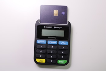 A small electronic card reader, featuring a keypad with numbers and function buttons. A card is inserted into the top slot, exposing its chip and indicating contactless functionality. The device has branded labels and a display screen.