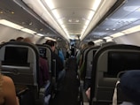 The interior of an airplane cabin with multiple rows of seats occupied by passengers. Overhead compartments are visible, and individual screens are mounted on the seat backs. The lighting is soft, and the cabin has a clean and organized appearance.