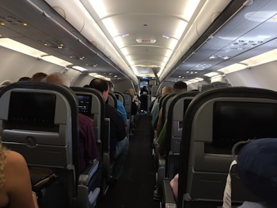 The interior of an airplane cabin with multiple rows of seats occupied by passengers. Overhead compartments are visible, and individual screens are mounted on the seat backs. The lighting is soft, and the cabin has a clean and organized appearance.