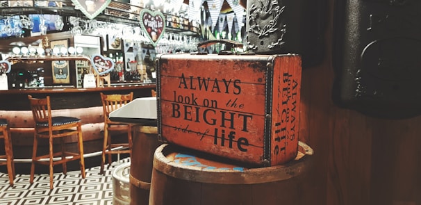 A vintage-style bar interior features a wooden counter with high chairs and glassware displayed on rows of shelves. An old suitcase with inspirational text is set atop a barrel, adding a retro charm to the scene. The floor has a geometric black and white tile pattern, and the walls are adorned with various signs and flags, enhancing the cozy and nostalgic atmosphere.