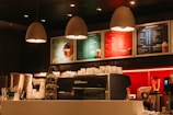 Warmly lit coffee shop interior with a modern espresso machine, stacks of takeaway cups, and colorful menu boards on the wall. Three pendant lamps hang above the counter, creating a cozy atmosphere.