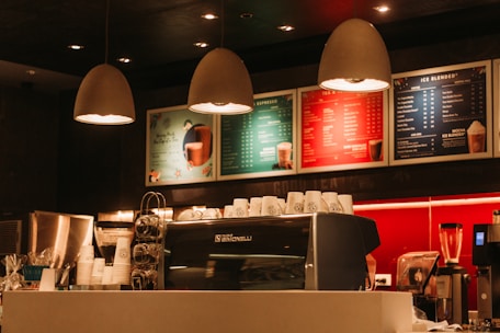 Warmly lit coffee shop interior with a modern espresso machine, stacks of takeaway cups, and colorful menu boards on the wall. Three pendant lamps hang above the counter, creating a cozy atmosphere.