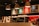 Warmly lit coffee shop interior with a modern espresso machine, stacks of takeaway cups, and colorful menu boards on the wall. Three pendant lamps hang above the counter, creating a cozy atmosphere.