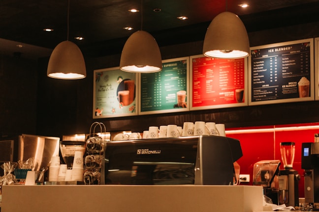 Warmly lit coffee shop interior with a modern espresso machine, stacks of takeaway cups, and colorful menu boards on the wall. Three pendant lamps hang above the counter, creating a cozy atmosphere.