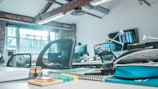 A modern office environment includes multiple workstations equipped with computers and office supplies. The atmosphere is enhanced with natural light streaming through large windows. The workspace features ergonomic chairs, desks with various items such as notebooks, pens, and a pencil case. Brick walls and exposed beams add an industrial aesthetic.