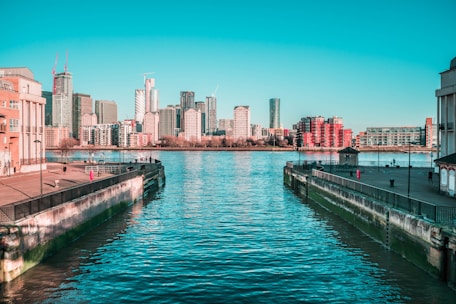A clear waterway, possibly a canal or river, is flanked by concrete walls leading towards a calm water surface. In the background, there is a cityscape with numerous modern high-rise buildings, some of which are still under construction with visible cranes. The sky is bright and clear, and there are people walking along the walkway on both sides of the water.