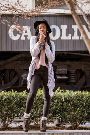 A young woman stands confidently on a low platform in front of a train car with the partially visible text 'CAROLIN'. She is dressed in a stylish, casual outfit consisting of a black hat, gray cardigan over a pink top, and black jeans. Her hands are near her face, and her expression is thoughtful. The background shows a mix of greenery and the train's metal components, while the foreground includes branches with sparse leaves.