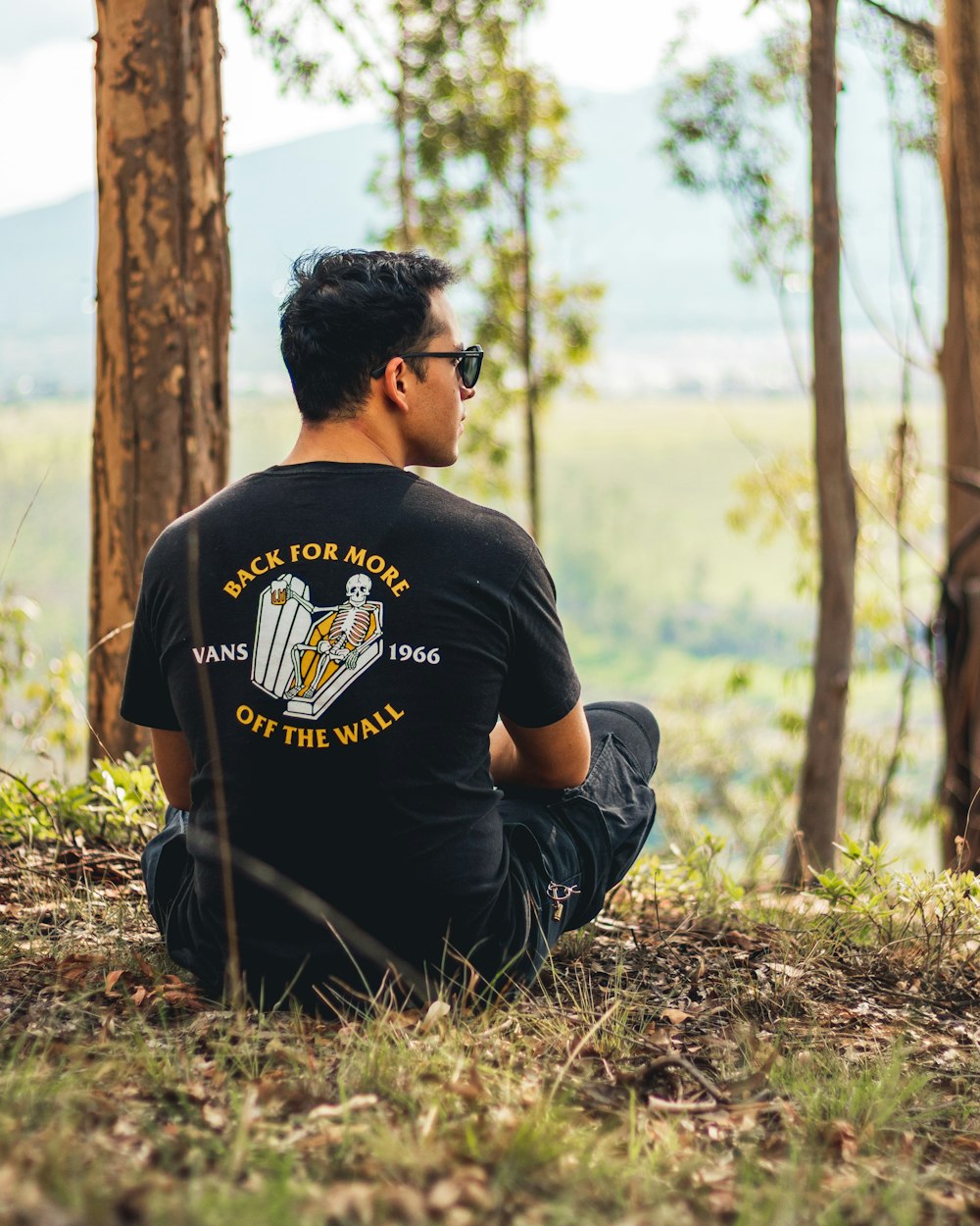 man in black crew neck t-shirt sitting on ground beside brown tree during daytime