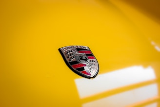 A close-up image of a car emblem featuring a shield with intricate designs in black, red, and silver against a glossy yellow background.