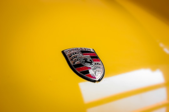 A close-up image of a car emblem featuring a shield with intricate designs in black, red, and silver against a glossy yellow background.