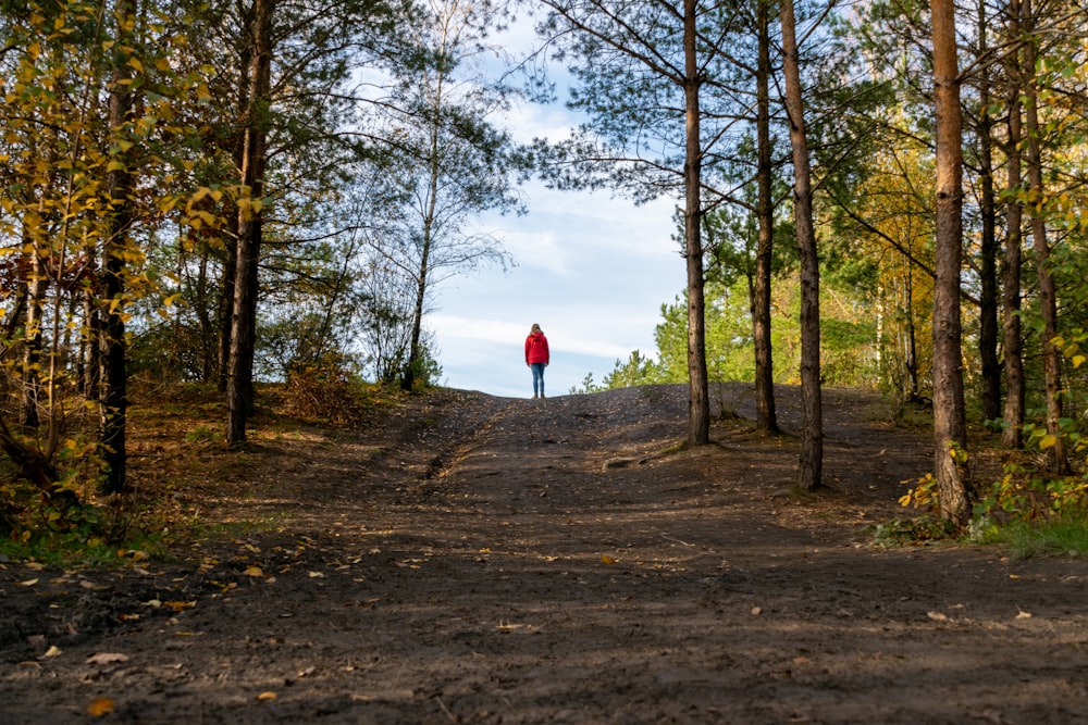 person in red jacket standing on brown dirt road between trees during daytime