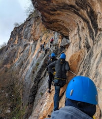 A group of people wearing blue helmets and harnesses climb along a narrow pathway on a steep rocky cliff. The surface of the cliff varies in color, with shades of brown, gray, and beige. Sparse vegetation is visible, indicating a rugged, natural environment.