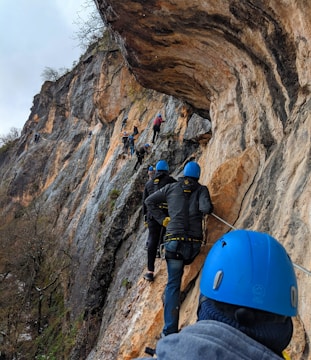 A group of people wearing blue helmets and harnesses climb along a narrow pathway on a steep rocky cliff. The surface of the cliff varies in color, with shades of brown, gray, and beige. Sparse vegetation is visible, indicating a rugged, natural environment.