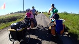 A family of four rides bicycles along a rural road. Two adults each have a child on the back of their bikes, with trailers attached carrying luggage. The road is surrounded by fields and greenery under a clear blue sky.
