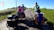 A family of four rides bicycles along a rural road. Two adults each have a child on the back of their bikes, with trailers attached carrying luggage. The road is surrounded by fields and greenery under a clear blue sky.