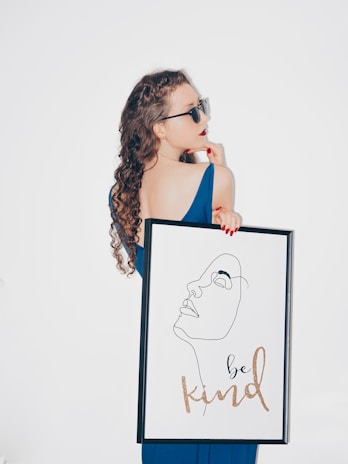 A woman with curly hair wearing sunglasses and a blue dress holds a framed artwork featuring a minimalist line drawing of a face with the words 'be kind' written below in cursive. The woman is gazing to the side with a thoughtful expression.