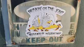 A sticker is placed on a weathered surface, featuring a cartoon drawing of two beer kegs with legs, wearing hats, appearing to be drunk and surrounded by cheese, pepperoni slices, and red cups. The text above reads 'DRINKIN' ON THE JOB!!!' with additional text 'NASTY NECKFACE!!! PIZZANISTA!!!'. The sticker overlays a fading 'KEEP OUT' sign.