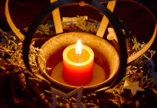 A single lit candle is placed in the center of a decorative arrangement with natural elements such as pine cones and twigs. The candle emits a warm, glowing light, creating a cozy and intimate atmosphere. Wooden star ornaments are visible around the arrangement, enhancing the festive and rustic aesthetic.