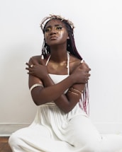 A woman with braided hair sits against a minimalistic white background. She wears a white dress and has her arms crossed over her chest. Her makeup is bold with golden eyeshadow and face accents. She wears a crown made of twigs and arm jewelry that adds to the visual contrast and artistic expression.