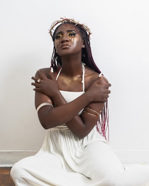A woman with braided hair sits against a minimalistic white background. She wears a white dress and has her arms crossed over her chest. Her makeup is bold with golden eyeshadow and face accents. She wears a crown made of twigs and arm jewelry that adds to the visual contrast and artistic expression.