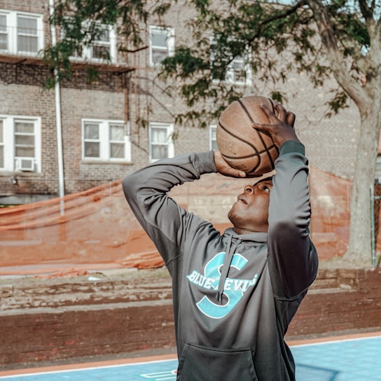 A person is preparing to shoot a basketball on an outdoor court surrounded by brick buildings and trees. The individual is wearing a gray hoodie with a large letter 'S' and the words 'Blue Devils' printed on it. The basketball court features a blue surface.