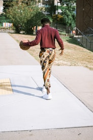 A person dressed in a maroon shirt and colorful patterned pants is walking down a sidewalk while carrying a basketball under one arm. The setting appears to be urban, with buildings and greenery visible in the background. The individual is wearing white sneakers.
