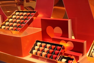 An assortment of chocolates shaped like hearts, arranged in gold-framed boxes. The chocolates vary in color, predominantly featuring shades of red and white. The background includes red packaging with heart designs that complement the chocolates, giving a romantic and thematic presentation.