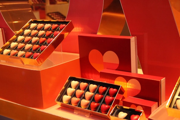 An assortment of chocolates shaped like hearts, arranged in gold-framed boxes. The chocolates vary in color, predominantly featuring shades of red and white. The background includes red packaging with heart designs that complement the chocolates, giving a romantic and thematic presentation.