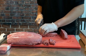 A person wearing gloves is slicing a large piece of raw red meat on a red cutting board. A brick wall forms the background, and part of the workspace includes a shiny metal surface.