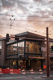 A modern, urban building located at a street intersection. The building has large glass windows and a sign that indicates a retail store. There are street signs, traffic lights, and overhead utility wires. The Washington street sign is visible. Brightly colored road barriers and construction signs are positioned on the street, indicating ongoing roadwork.