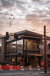 A modern, urban building located at a street intersection. The building has large glass windows and a sign that indicates a retail store. There are street signs, traffic lights, and overhead utility wires. The Washington street sign is visible. Brightly colored road barriers and construction signs are positioned on the street, indicating ongoing roadwork.