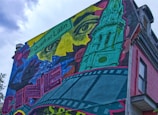 A vibrant mural covers the side of a building, featuring a mix of artistic elements. The design includes a large, abstract face with intense eyes and a green architectural structure resembling a tower. There is a blend of film strip motifs and colorful geometric shapes, with a sign labeled 'Quartier Latin'.