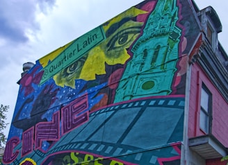 A vibrant mural covers the side of a building, featuring a mix of artistic elements. The design includes a large, abstract face with intense eyes and a green architectural structure resembling a tower. There is a blend of film strip motifs and colorful geometric shapes, with a sign labeled 'Quartier Latin'.