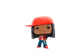 A vinyl figurine with a cartoonish style stands against a white background. It has dark skin, long black hair, and is wearing a red cap with the brim to the side, a matching red jacket, blue pants, and red shoes.