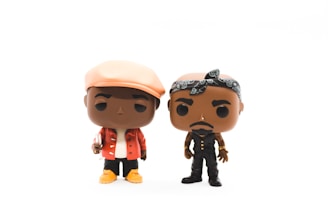 Two stylized vinyl figures are depicted, each with distinct outfits and accessories. The figure on the left is wearing a light-colored hat, a red jacket, white shirt, and yellow shoes, while holding a small item. The figure on the right has a black bandana, black vest, and pants, with gold accessories.