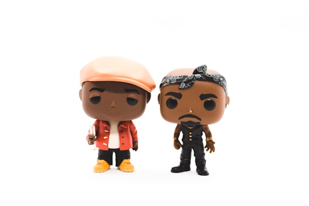 Two stylized vinyl figures are depicted, each with distinct outfits and accessories. The figure on the left is wearing a light-colored hat, a red jacket, white shirt, and yellow shoes, while holding a small item. The figure on the right has a black bandana, black vest, and pants, with gold accessories.