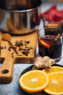 A cozy setup featuring a wooden cutting board with various spices scattered on it, next to a stainless steel pot. A glass of dark-colored beverage with a slice of orange and a cinnamon stick is placed nearby. In the foreground, slices of orange and a piece of ginger are arranged on a small plate.