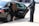 A person in a black suit stands by an open car door, assisting a person with long hair and a black dress into the back seat of a black sedan parked on a sidewalk. The setting appears to be an urban street.