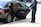 A person in a black suit stands by an open car door, assisting a person with long hair and a black dress into the back seat of a black sedan parked on a sidewalk. The setting appears to be an urban street.