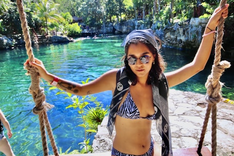 A woman in a zebra-patterned bikini and sunglasses stands holding onto two thick ropes by a clear blue-green water body. She is wearing a printed scarf on her head and has a tattoo on her arm. The background features lush greenery and rocky formations surrounding the water.