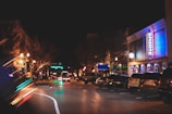 A nighttime urban street scene with several parked cars lining the side of the road. Buildings on the right feature bright neon lights and signs, including one that reads 'Puckett's.' The street is illuminated by overhead street lamps and the glow from the buildings, creating a lively, bustling atmosphere.