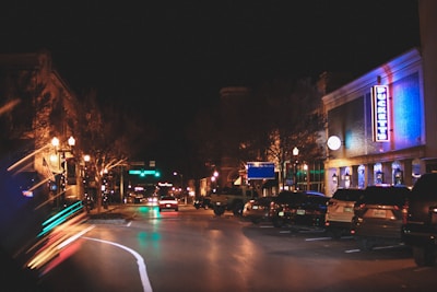 A nighttime urban street scene with several parked cars lining the side of the road. Buildings on the right feature bright neon lights and signs, including one that reads 'Puckett's.' The street is illuminated by overhead street lamps and the glow from the buildings, creating a lively, bustling atmosphere.