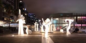 A series of illuminated abstract sculptures are arranged in an outdoor plaza. The sculptures vary in shape and size, exhibiting a modern architectural style. Surrounding the art, urban buildings and a glass-fronted structure with colorful lights are visible, contributing to a contemporary urban landscape. The scene is set at night, creating a captivating juxtaposition with the glowing sculptures.