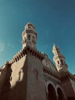 An ornate building showcases intricate architectural details with two tall minarets. The structure features detailed stone carvings and arched windows, influenced by Islamic architectural style.