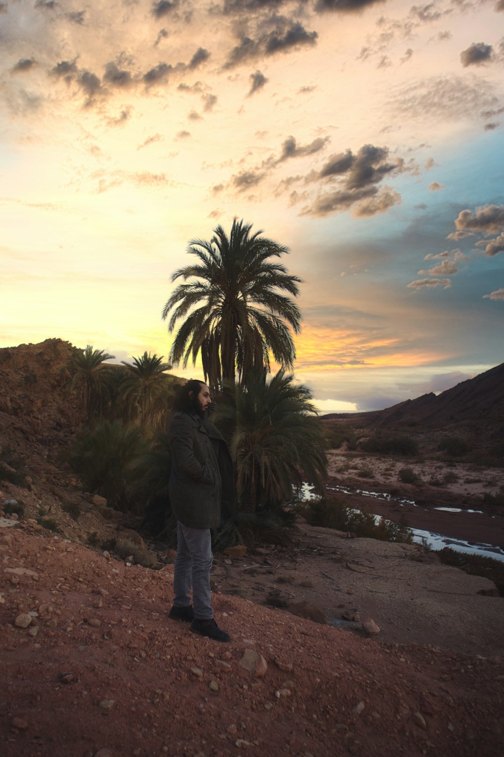 man in black jacket standing on brown sand near body of water during sunset